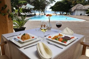 A table set with plates of food, a wine glass, and a vase with flowers, overlooking a pool and beach.