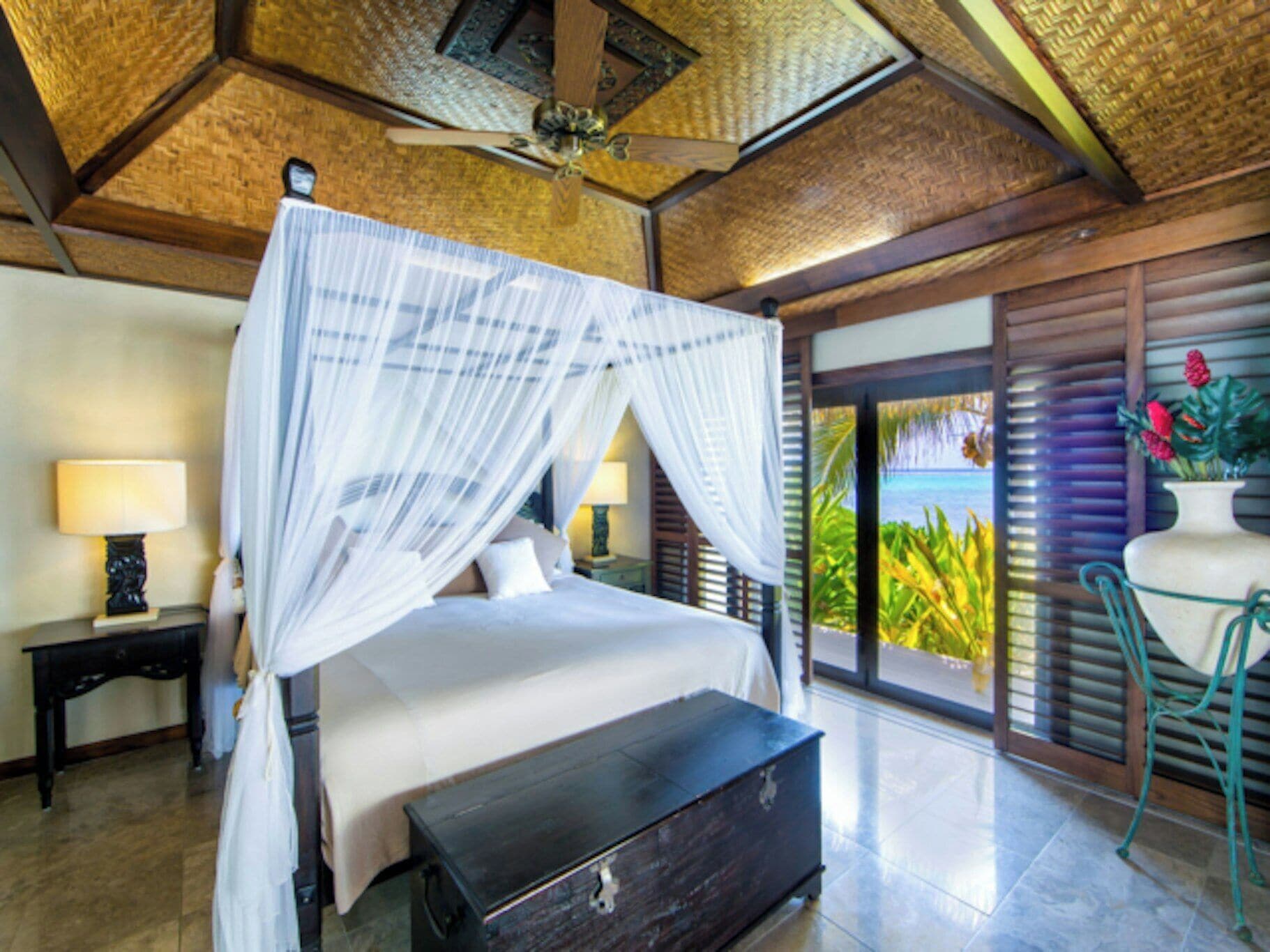 A four-poster bed with a canopy, a bedside table with a lamp, a bench, and a view of the ocean through a sliding door.