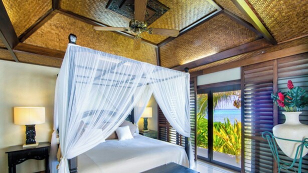 A four-poster bed with a canopy, a bedside table with a lamp, a bench, and a view of the ocean through a sliding door. - Te Manava Luxury Villas Spa (Rarotonga)