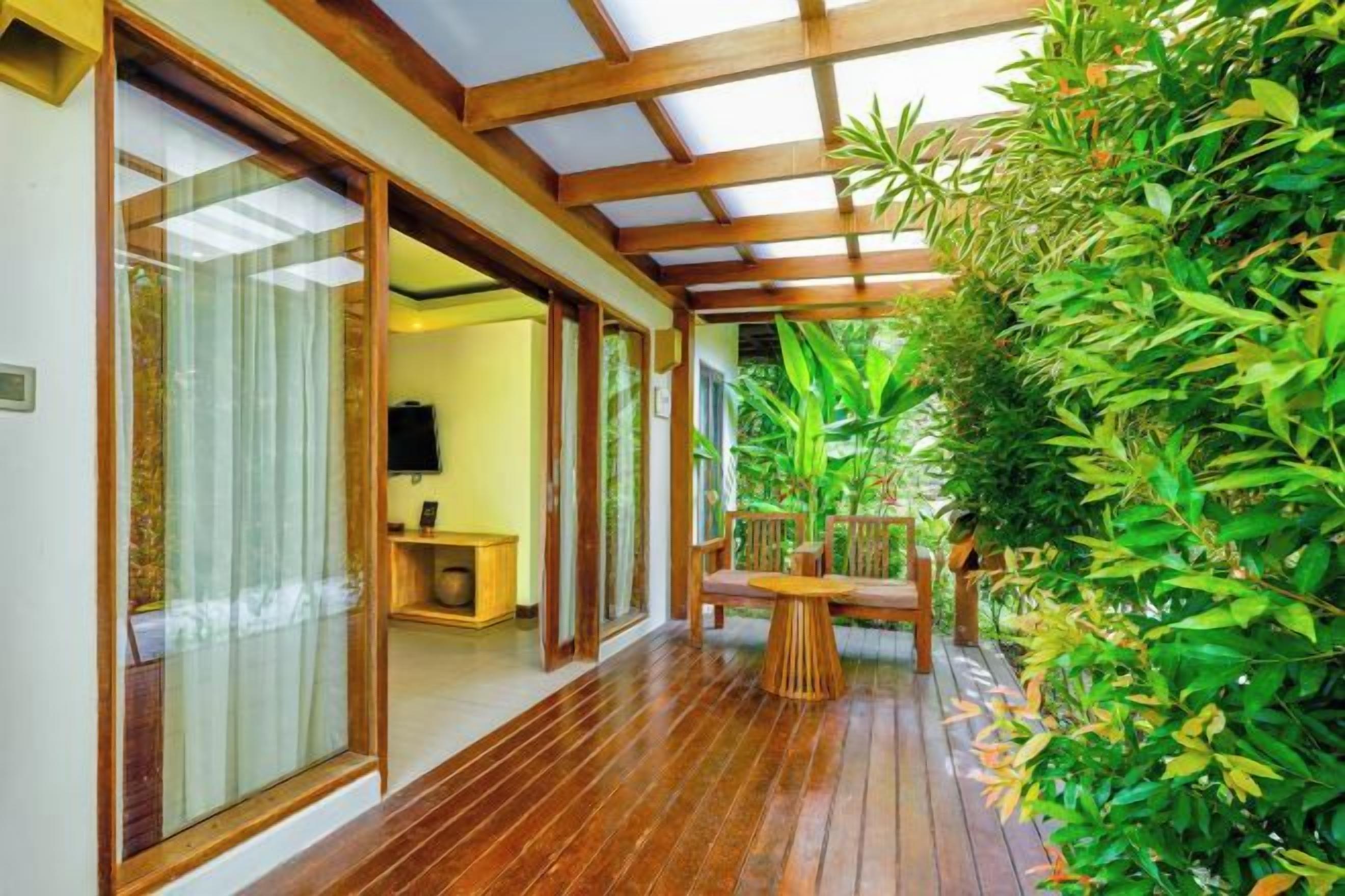 A wooden deck with a glass door leading to a room, surrounded by lush greenery.