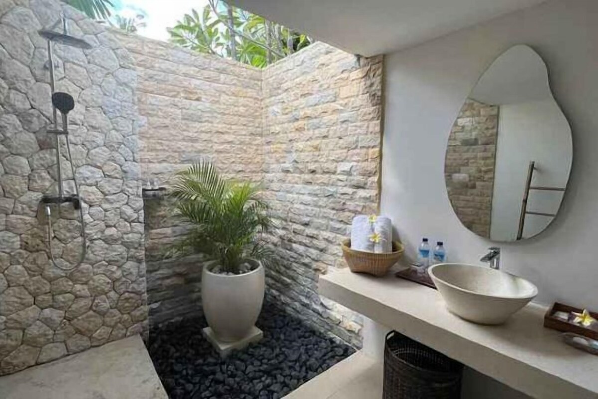 Un baño con pared de piedra, una planta en maceta, un espejo grande y un lavabo con artículos de aseo.