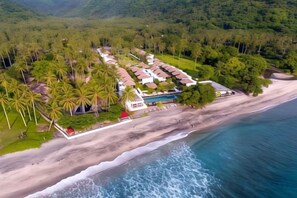 Vista aérea de un resort con alojamiento frente a la playa, piscina y exuberante vegetación.