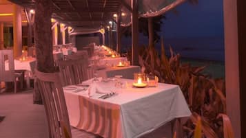 An outdoor dining area with tables set for a meal, surrounded by chairs and palm plants.