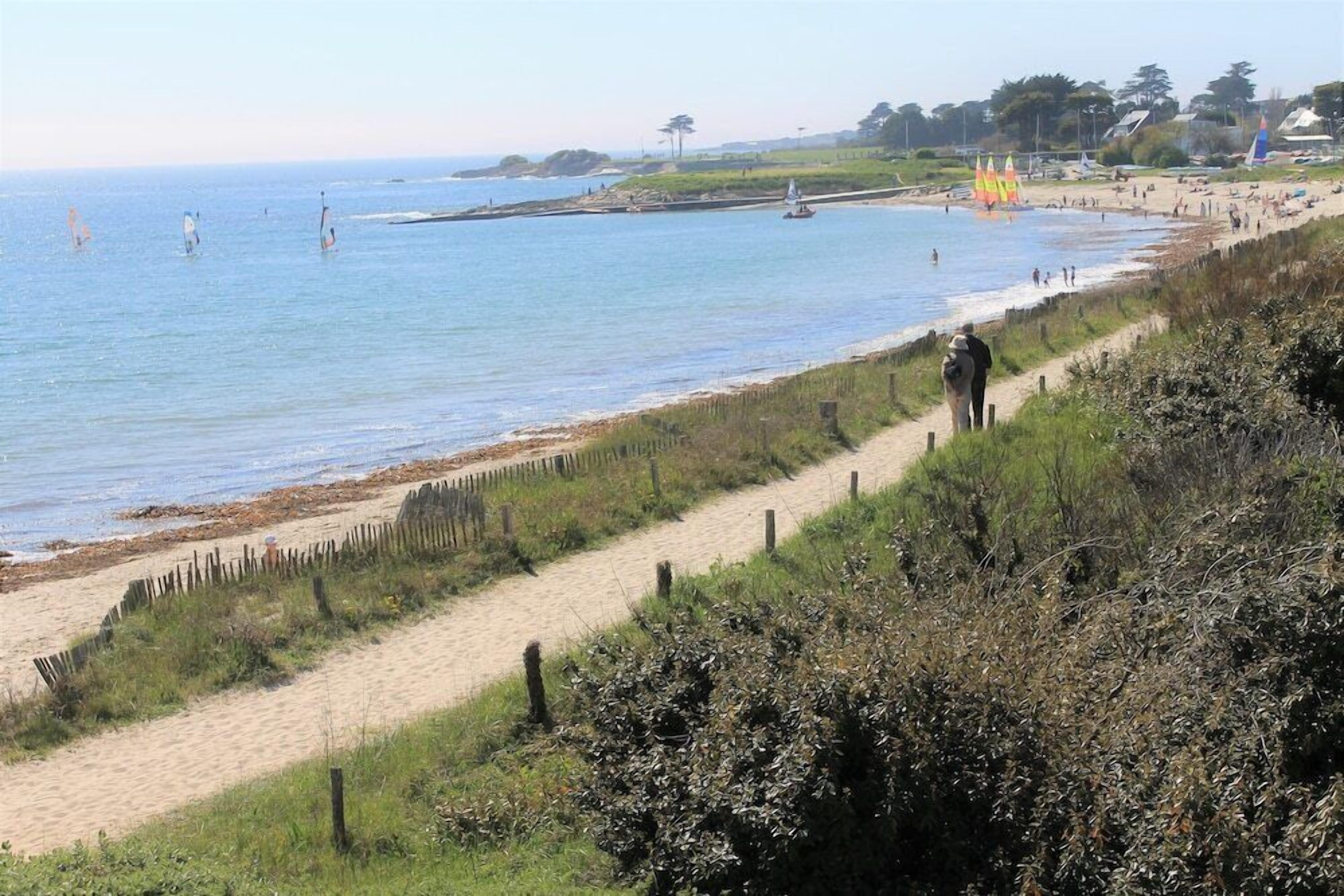 A coastal beach with a sandy path leading to the water, a fence along the edge, and a few people in the distance.