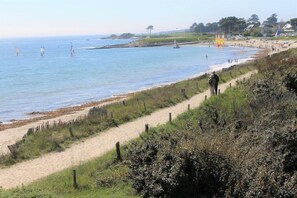 Una playa costera con un sendero arenoso que lleva al agua, una cerca a lo largo de la orilla y algunas personas a lo lejos.
