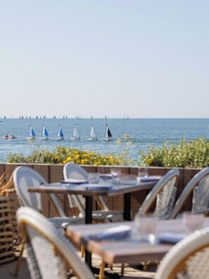 Un área de comedor frente al mar con sillas y mesas de mimbre, con vista a un mar tranquilo y veleros.