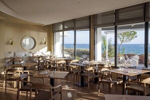 Ein Restaurant mit Tischen, Stühlen, großen Fenstern und Meerblick.