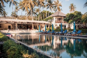 A pool area with lounge chairs and umbrellas, surrounded by palm trees and a building with a thatched roof. - Living Asia Resort Spa Lombok (Senggigi)