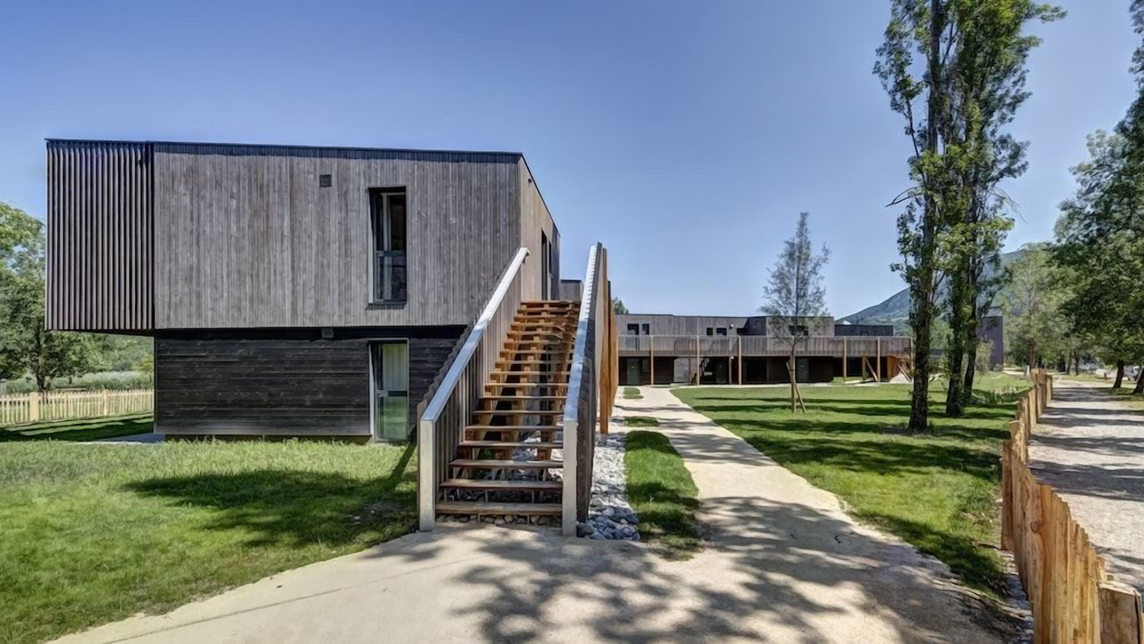 A modern building with a wooden exterior, a staircase leading to a door, and a pathway with trees.
