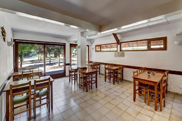 A dining area with multiple wooden tables and chairs, large windows with blinds, and a view of the outdoors.