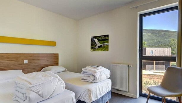 A hotel room with two beds, a chair, a window with a view of a building and hills, and a picture on the wall.