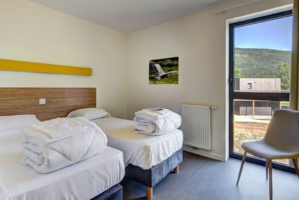 A hotel room with two beds, a chair, a window with a view of a building and hills, and a picture on the wall.