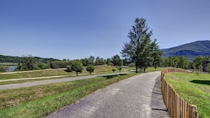 A paved pathway with a wooden fence on one side, surrounded by grassy areas and trees, leading towards a body of water and hills in the distance. - Le Village des Oiseaux (Motz)