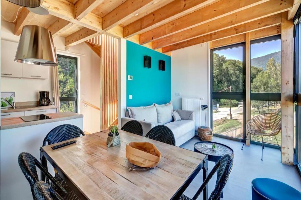 A modern interior with a wooden dining table, a sofa, and a view of the outdoors.