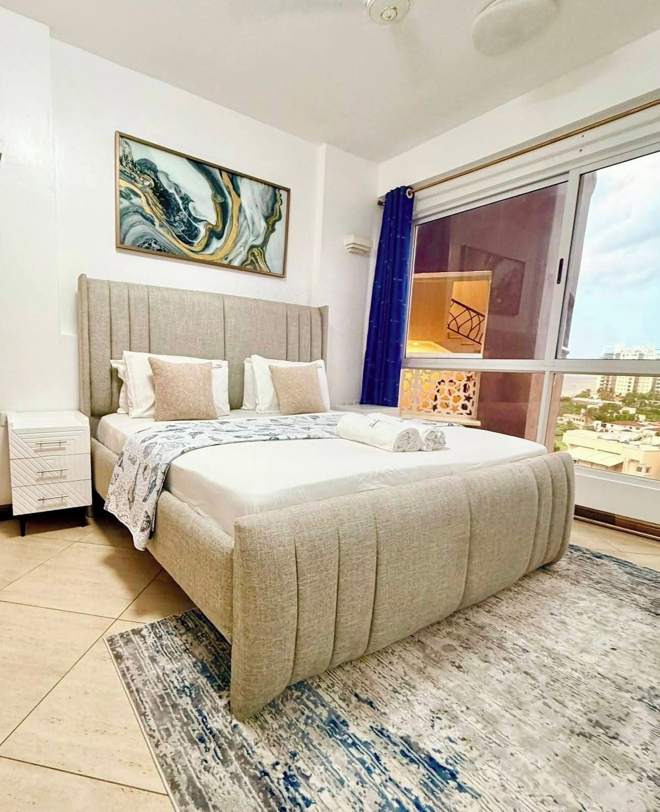 Un dormitorio moderno con una cama grande, una mesita de noche, una ventana con cortinas y vistas a la ciudad.