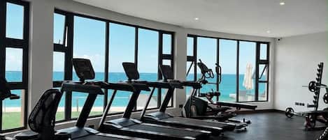 Une salle de sport équipée de tapis de course, de vélos d’exercice et d’appareils de musculation, avec de grandes fenêtres offrant une vue sur l’océan.