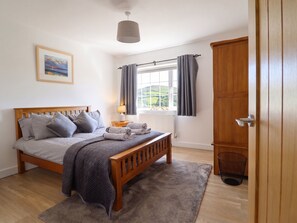 A bedroom with a wooden bed, grey bedding, a window with curtains, a radiator, and a painting on the wall. - Tyn Y Weirglodd (Caernarfon)