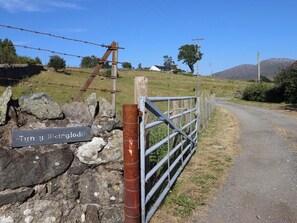 A rural road with a metal gate and a stone wall with a sign. - Tyn Y Weirglodd (Caernarfon)