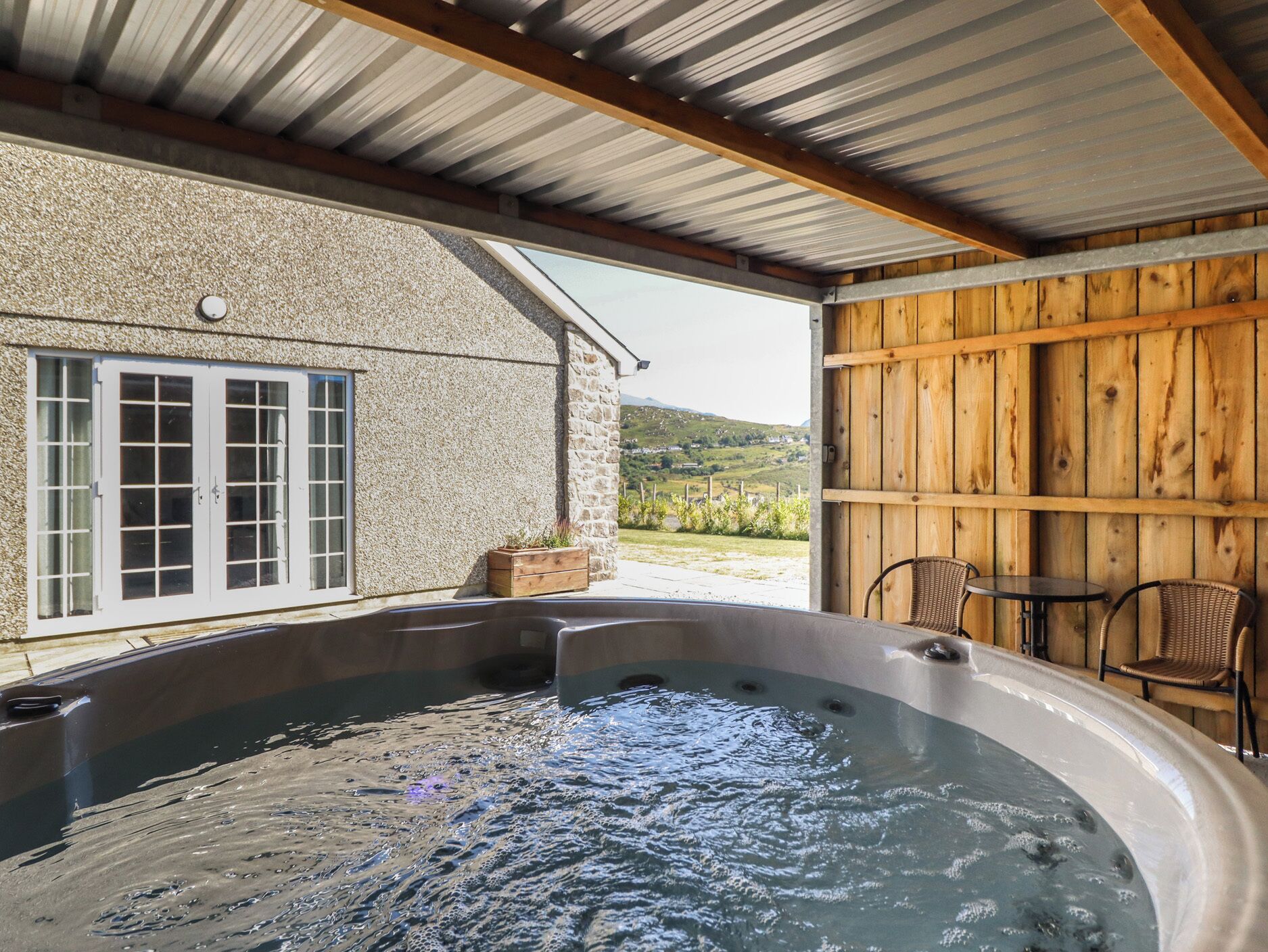 A hot tub area with a stone wall, wooden paneling, and a view of a green landscape.