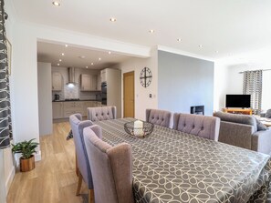 A modern dining area with a large table, multiple chairs, and a kitchen in the background. - Tyn Y Weirglodd (Caernarfon)