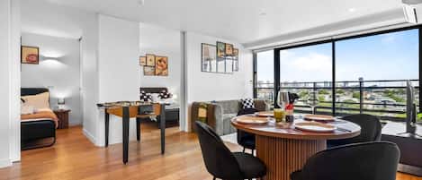 A modern living room with a dining area, a foosball table, and a large window offering a city view.