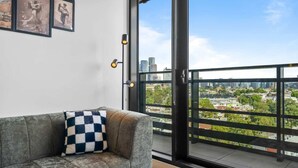 Ein graues Sofa mit einem karierten Kissen, Blick auf eine Stadtlandschaft durch ein großes Fenster und ein gerahmtes Bild an der Wand.