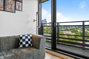 A grey sofa with a checkered pillow, a cityscape view through a large window, and a framed picture on the wall. - Walk to Melbourne Uni | Foosball & Free Parking (North Melbourne)
