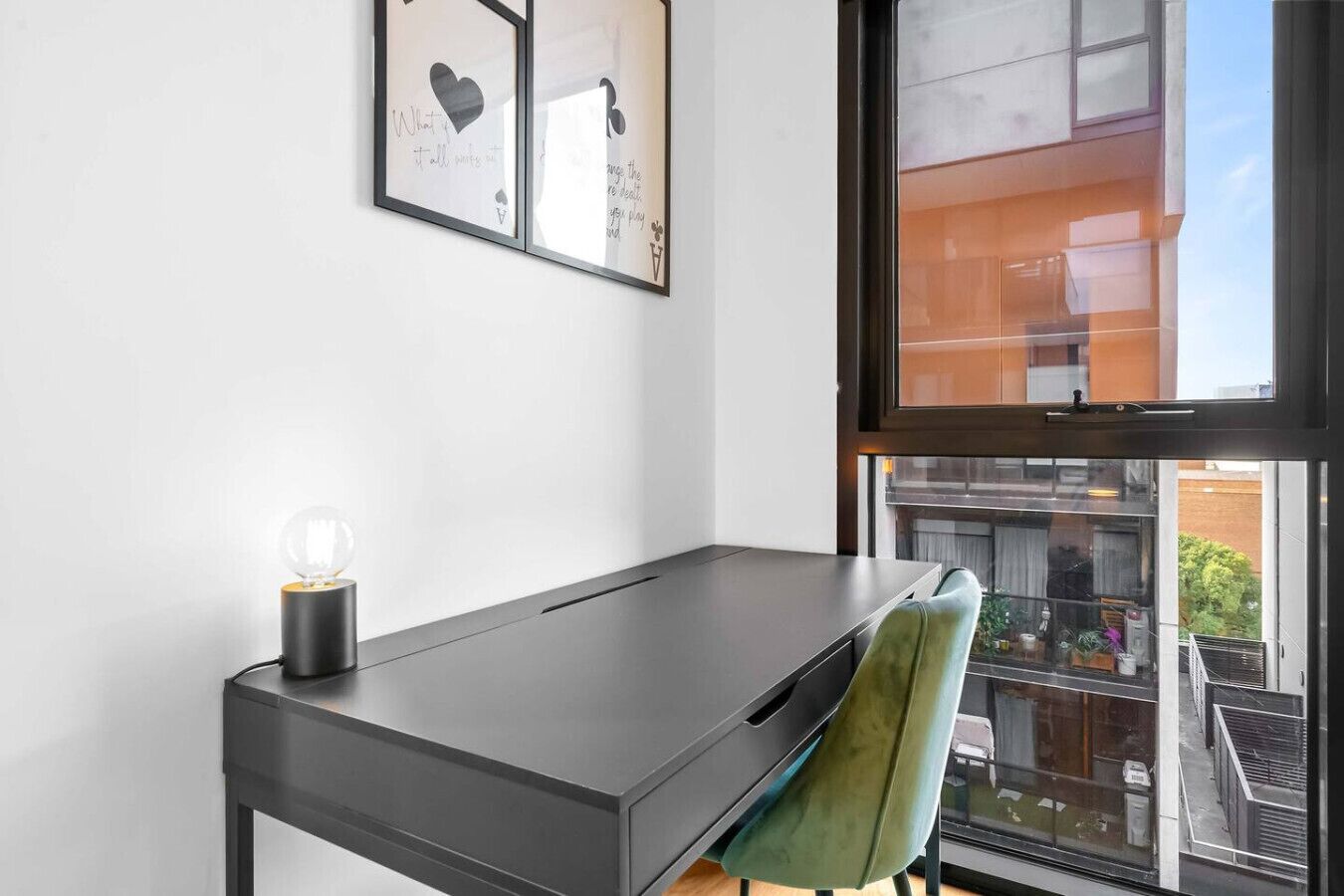 Une chambre moderne équipée d’un bureau, d’une chaise, d’une fenêtre donnant sur des immeubles et d’œuvres d’art encadrées accrochées au mur.