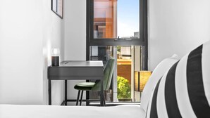 Ein kleines Zimmer mit einem Bett, einem Schreibtisch und einem Stuhl. Ein Fenster bietet Blick auf Gebäude und Grünflächen.