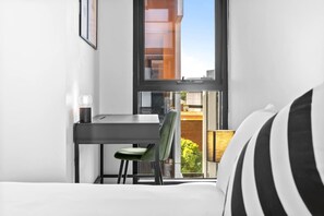 A small room with a bed, a desk, and a chair. There is a window with a view of buildings and greenery outside. - Walk to Melbourne Uni | Foosball & Free Parking (North Melbourne)