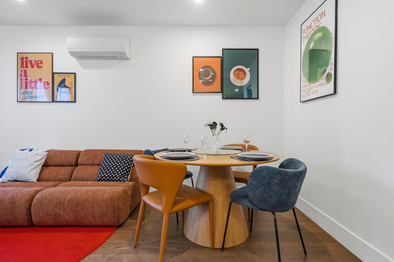 A modern living room with a brown sofa, orange chairs, a wooden dining table set for two, and a red rug.