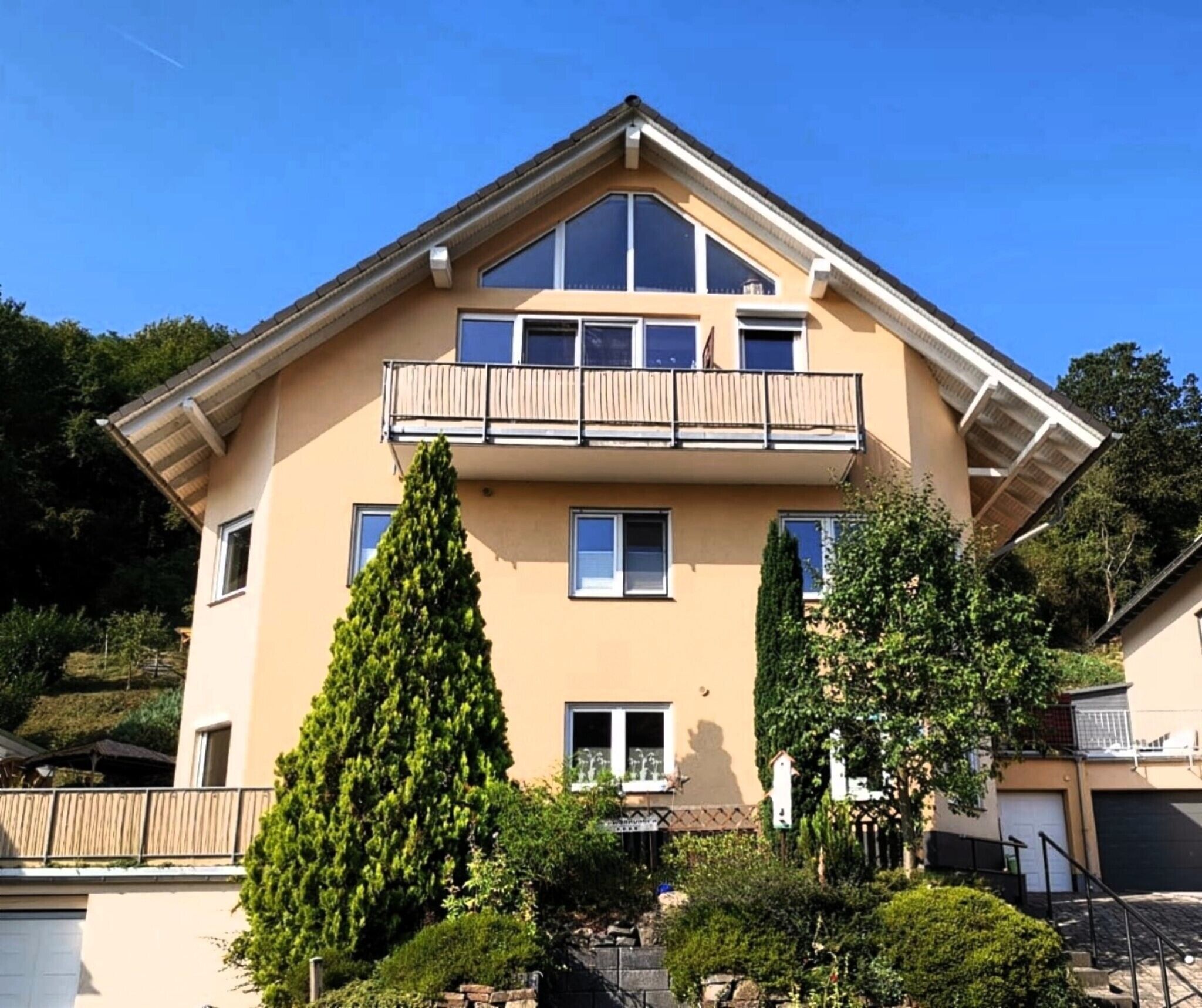 A two-story residential building with a balcony on the upper floor, surrounded by greenery.