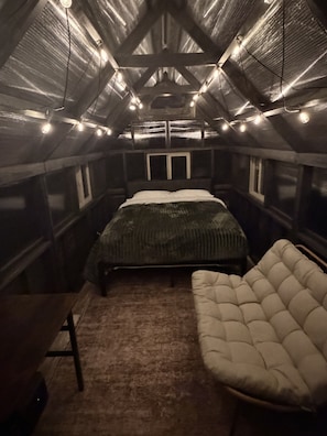 A small, dimly lit attic bedroom with a single bed, a cushioned chair, and a table. - Alderwood Polaris | Glamping Under the North Star (Wilson)