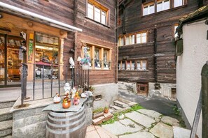 A narrow alleyway with wooden buildings, a small outdoor seating area with a barrel, and a shop with glass display windows. - Haus Kirchplatz - The Maisonnette (Zermatt)