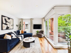 A modern living room with a dark blue sofa, a round coffee table, and a large window offering a view of the outdoors. - Westminster Residences by apt. (London)