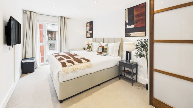 A modern hotel room with a large bed, a TV mounted on the wall, a bedside table with a lamp, and a view of the outside through a window. - Westminster Residences by apt. (London)