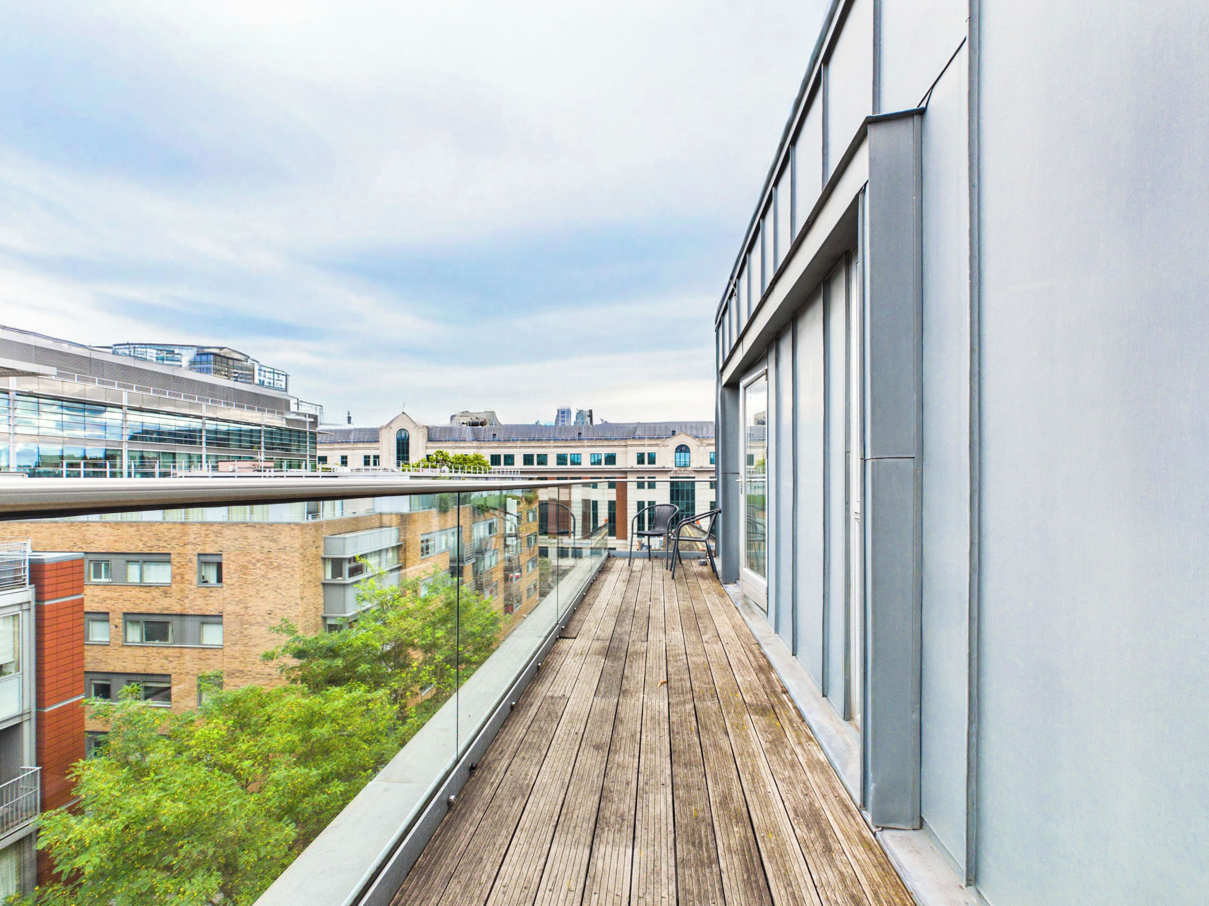 Un balcon doté d’un plancher en bois, offrant une vue sur un paysage urbain avec des bâtiments modernes et un ciel dégagé.