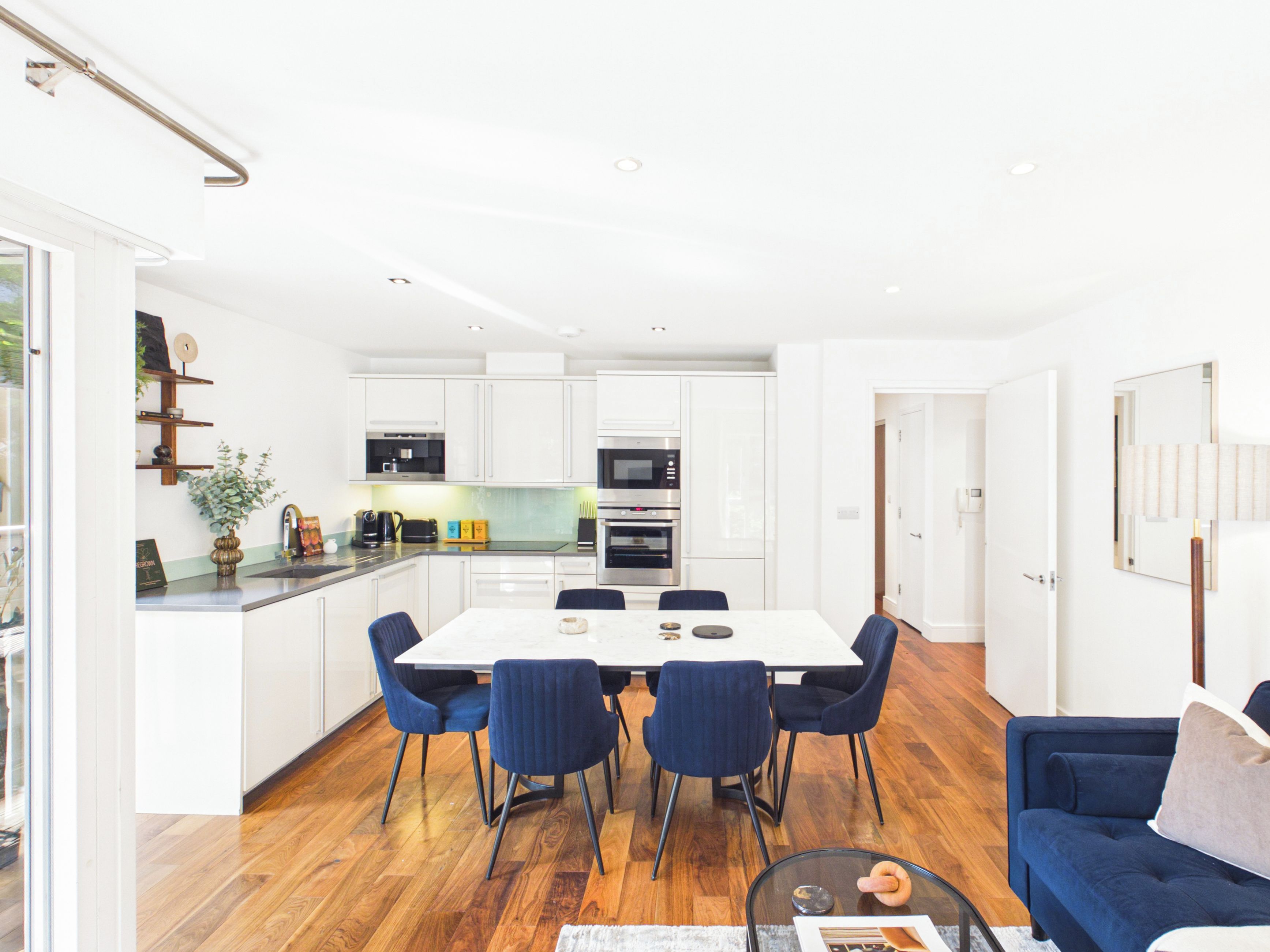 A modern kitchen with white cabinets, a dining area with a white table and blue chairs, and a living room with a blue sofa.