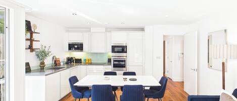 A modern kitchen with white cabinets, a dining area with a white table and blue chairs, and a living room with a blue sofa.