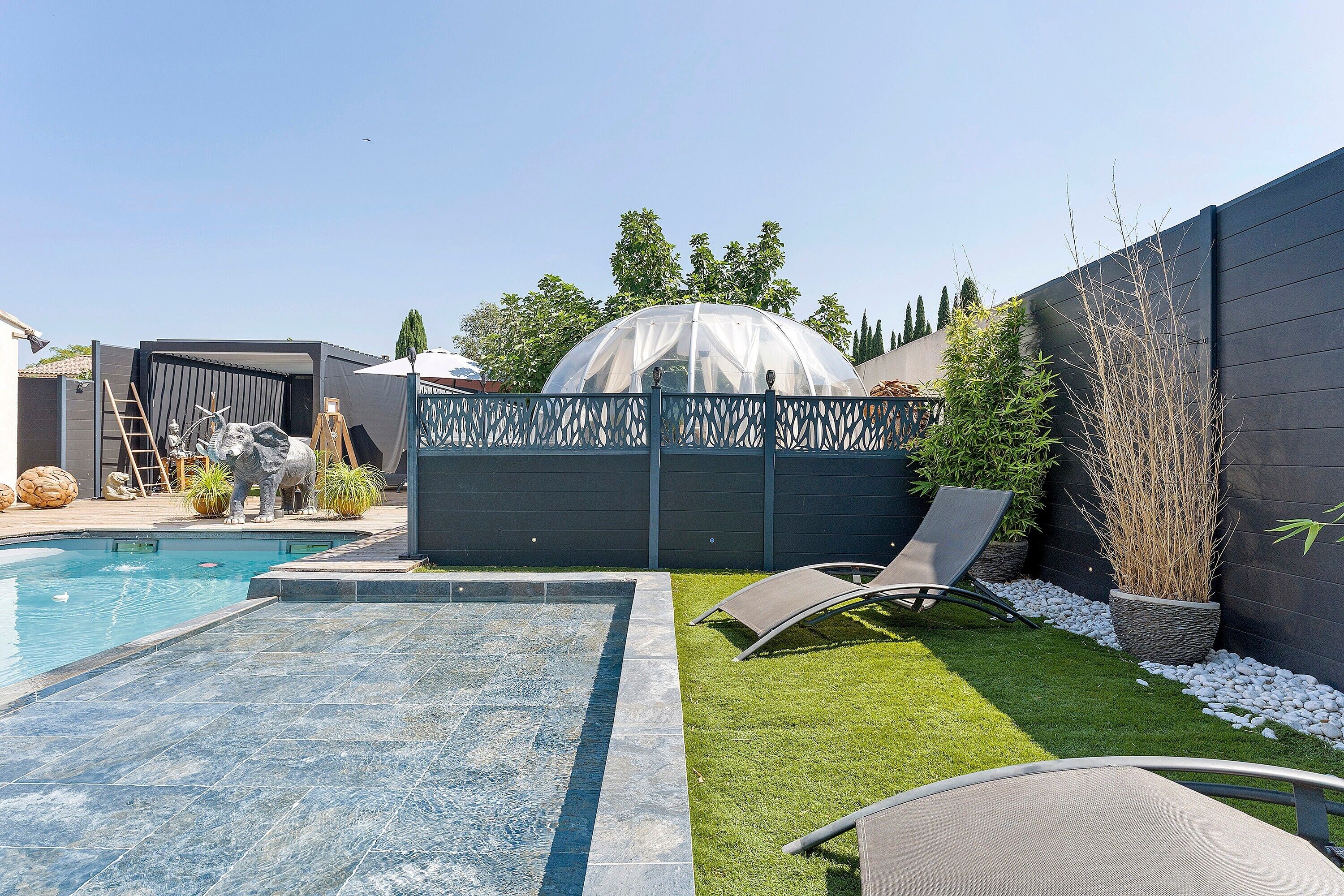 Ein Garten mit Swimmingpool, einem überdachten Gewächshaus und einer Sitzgruppe mit Liegestühlen.