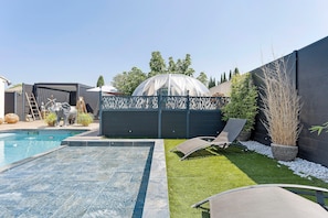 Ein Garten mit Swimmingpool, einem überdachten Gewächshaus und einer Sitzgruppe mit Liegestühlen.