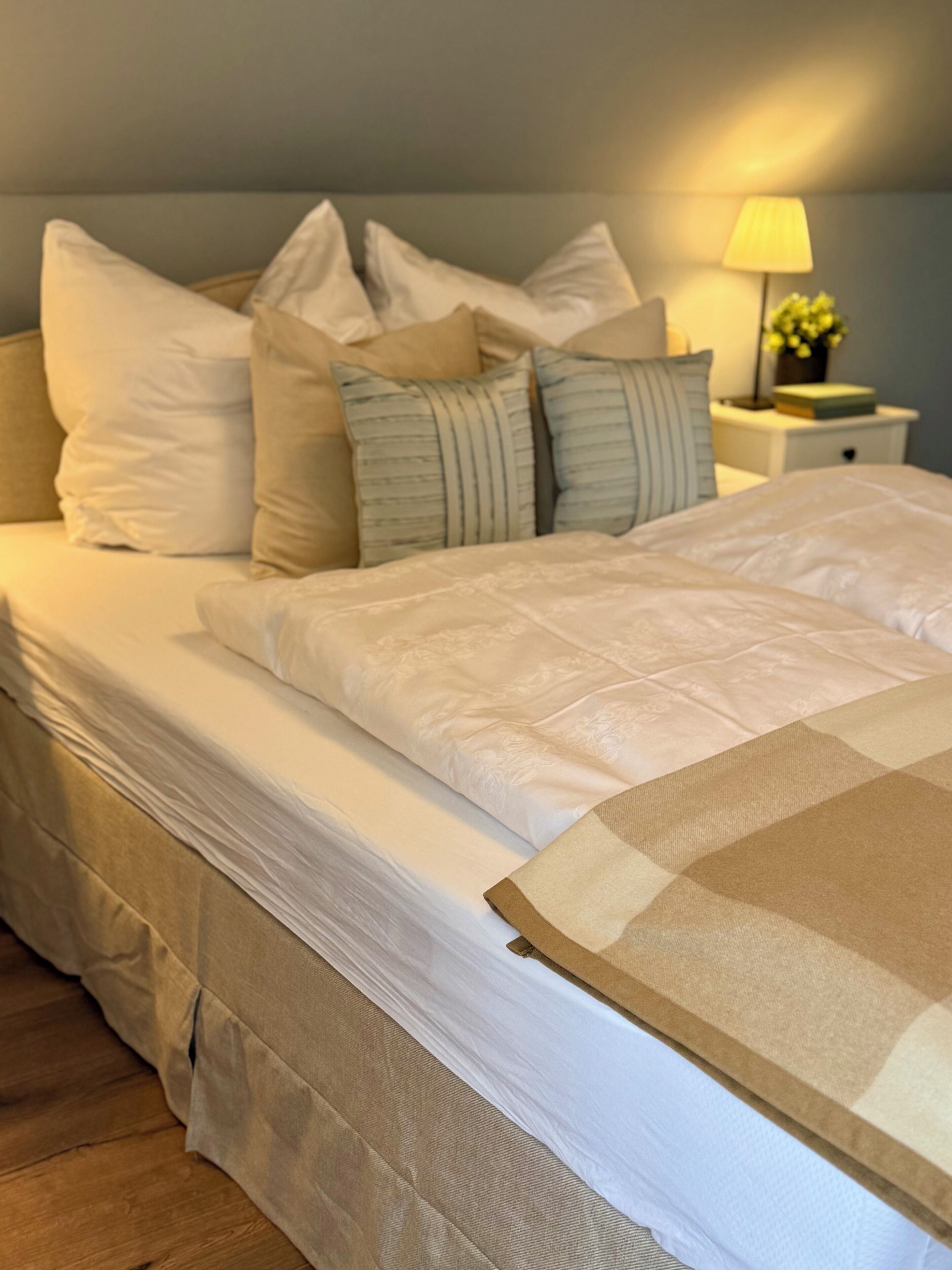 A neatly made bed with a white duvet and multiple pillows, a bedside table with a lamp, and a small plant.
