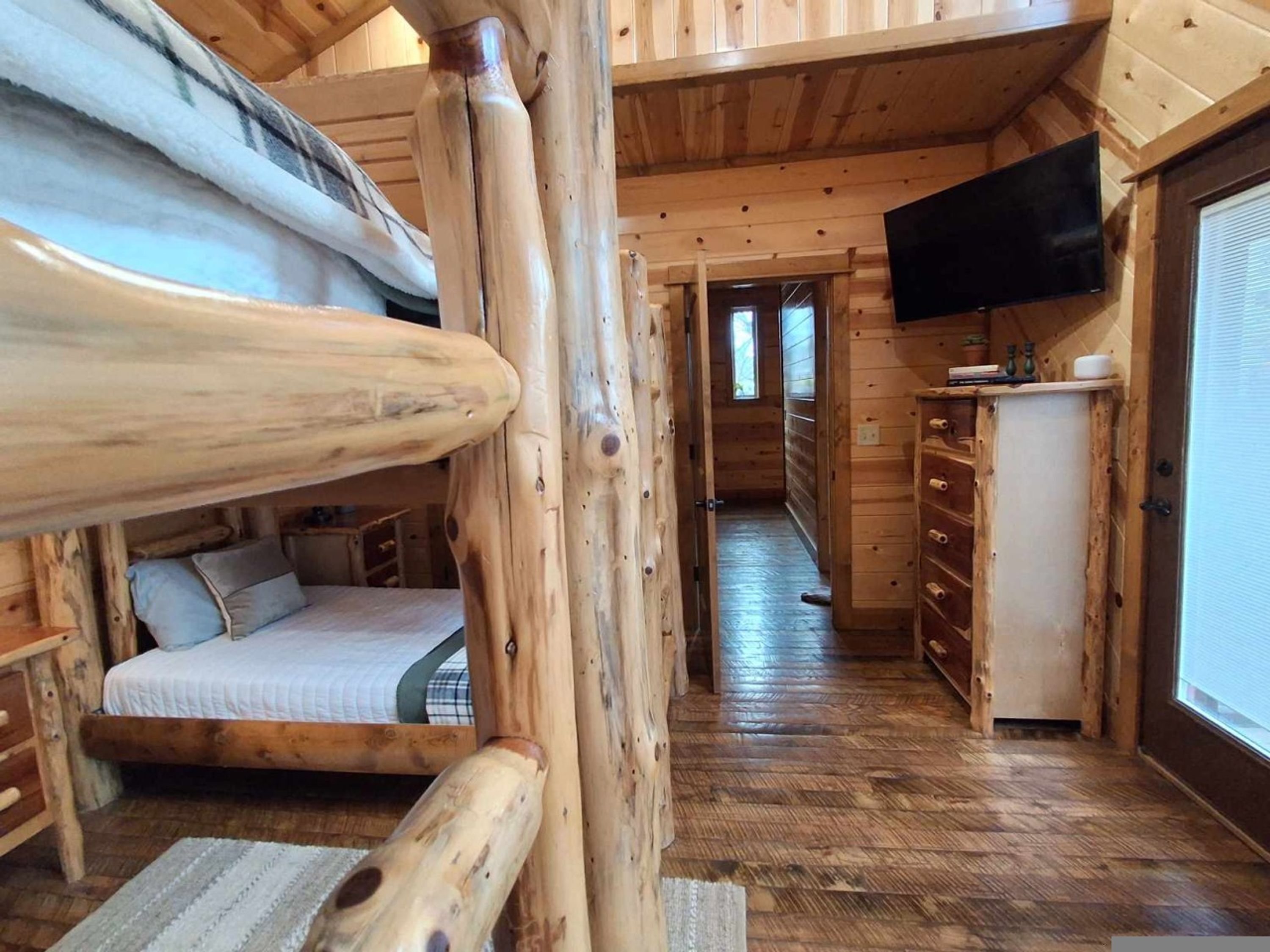 Interior de una cabaña de madera con una cama individual, una zona de cocina con televisor y un pasillo que conduce a otra habitación.