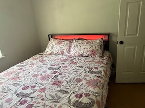 Ein Bett mit floralem Überwurf und Kissen, eine weiße Tür rechts und ein Fenster links.