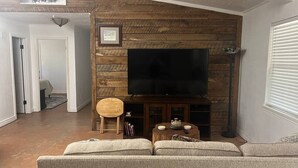 Sala de estar com televisão de tela plana fixada em uma parede de madeira, uma cadeira de madeira redonda, uma mesa pequena com objetos decorativos e um sofá.