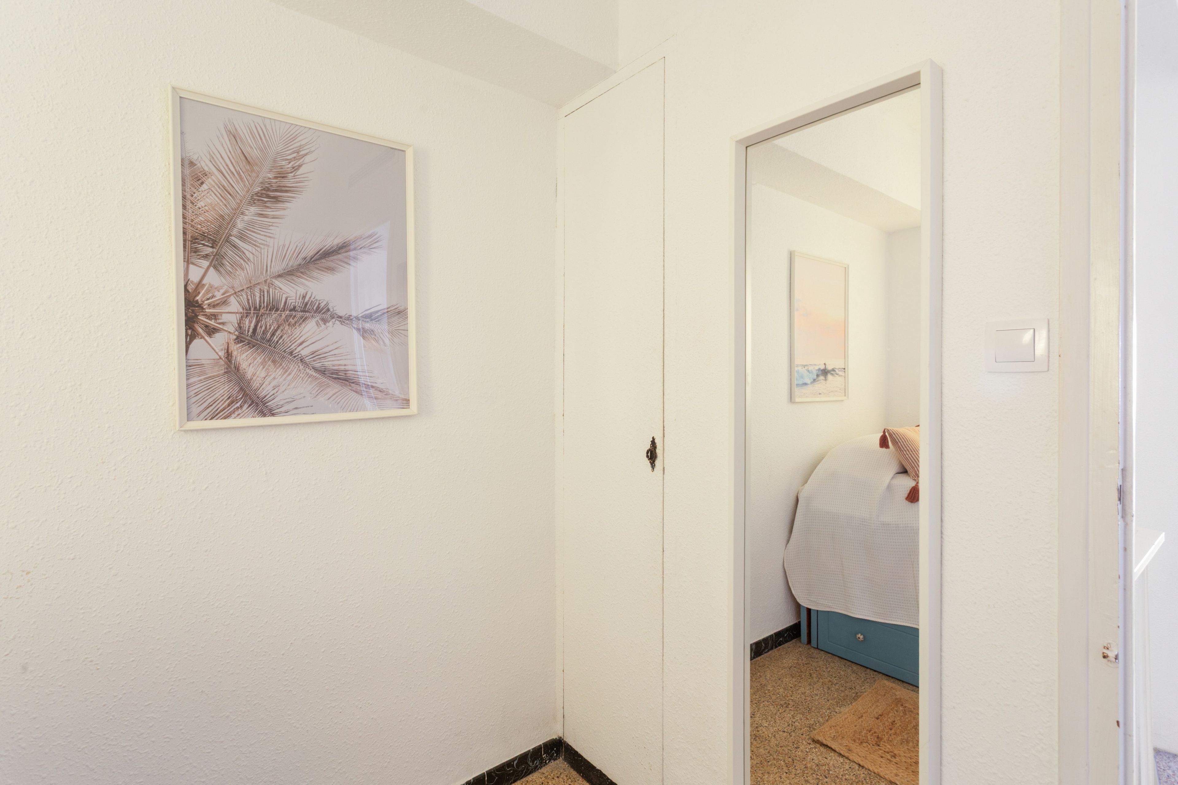 A hallway with a framed artwork on the wall, a door leading to a bedroom, and a visible bed with a blue headboard.