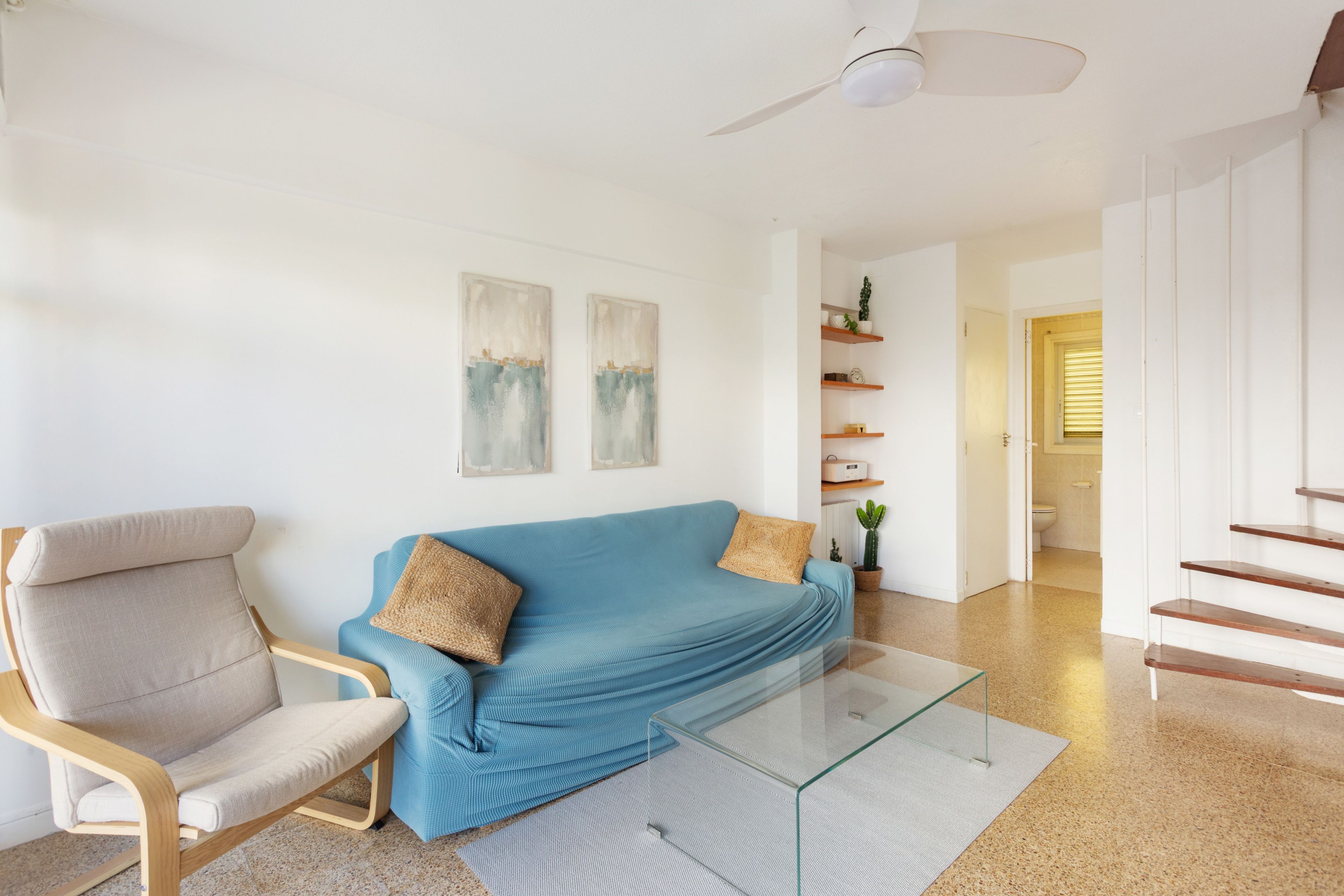 A modern living room with a blue sofa, a wooden chair, and a glass coffee table. There are two wall-mounted paintings, a ceiling fan, and a staircase leading to another room.