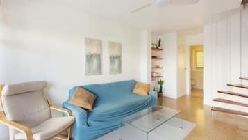 A modern living room with a blue sofa, a wooden chair, and a glass coffee table. There are two wall-mounted paintings, a ceiling fan, and a staircase leading to another room.