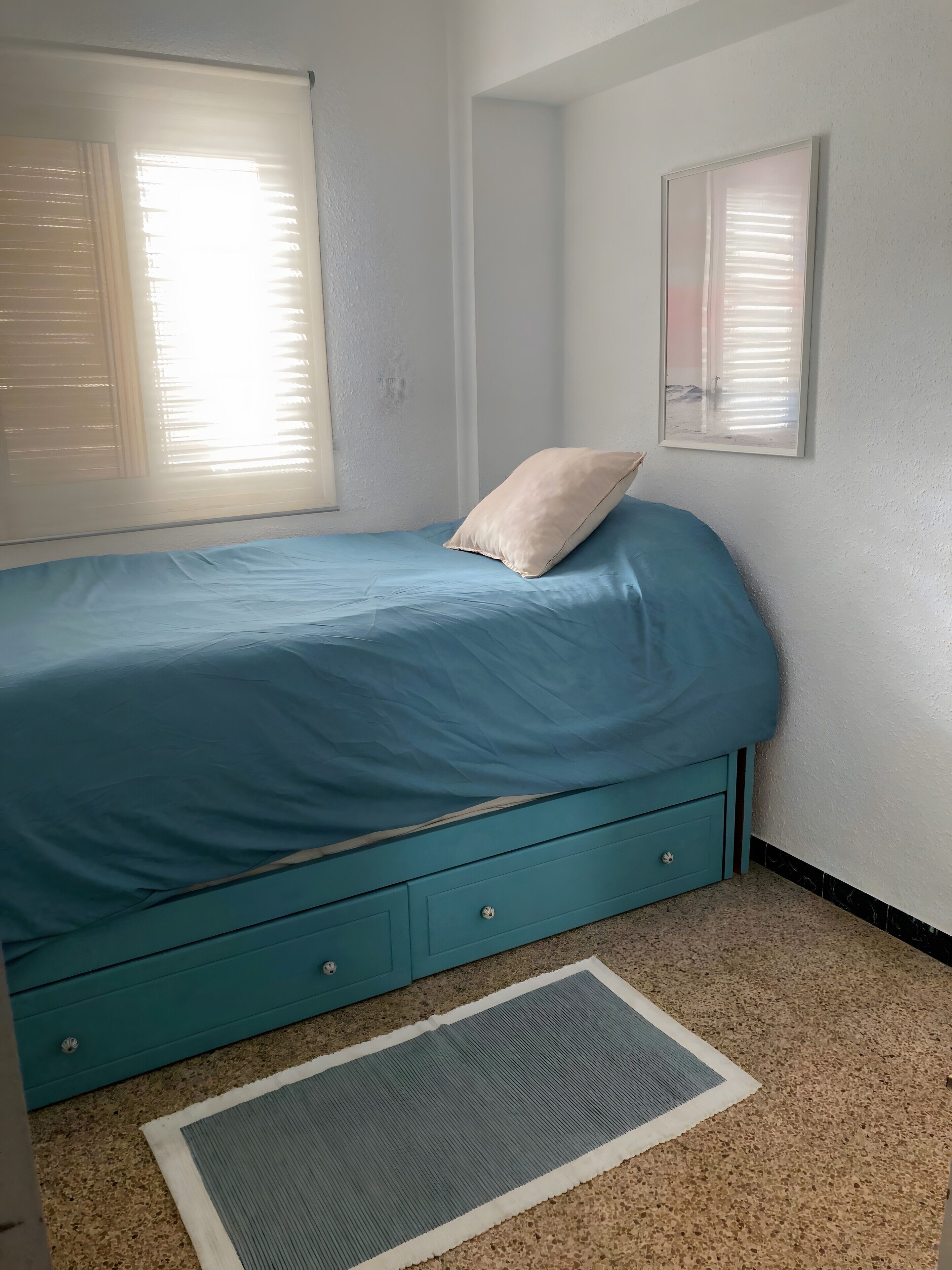 A bedroom with a bed, a window with blinds, a wall-mounted picture frame, and a small rug.