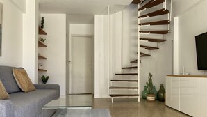 A modern living room with a grey sofa, a glass coffee table, a staircase with wooden steps, and a flat-screen TV on a white cabinet.